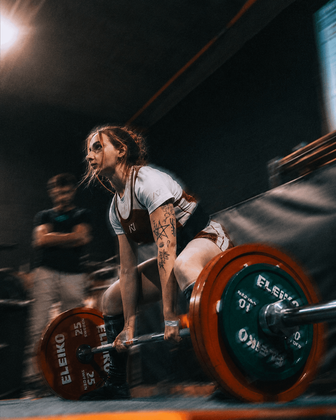 Powerlifting athlete lifting a barbell during a competition, with her coach watching